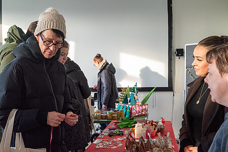 Adventsbasar Auw Mehrere Menschen stehen an einem Tisch mit handgemachten Weihnachtsartikeln. Im Vordergrund betrachtet eine ältere Person mit Mütze und Brille kleine Bastelarbeiten, während zwei weitere Personen hinter dem Tisch stehen und ins Gespräch vertieft sind. Auf dem Tisch liegen dekorative Gläser, Kerzen, Karten und kleine Weihnachtsbäume. Die Szene spielt in einem hellen Innenraum und vermittelt eine ruhige, vorweihnachtliche Marktatmosphäre.