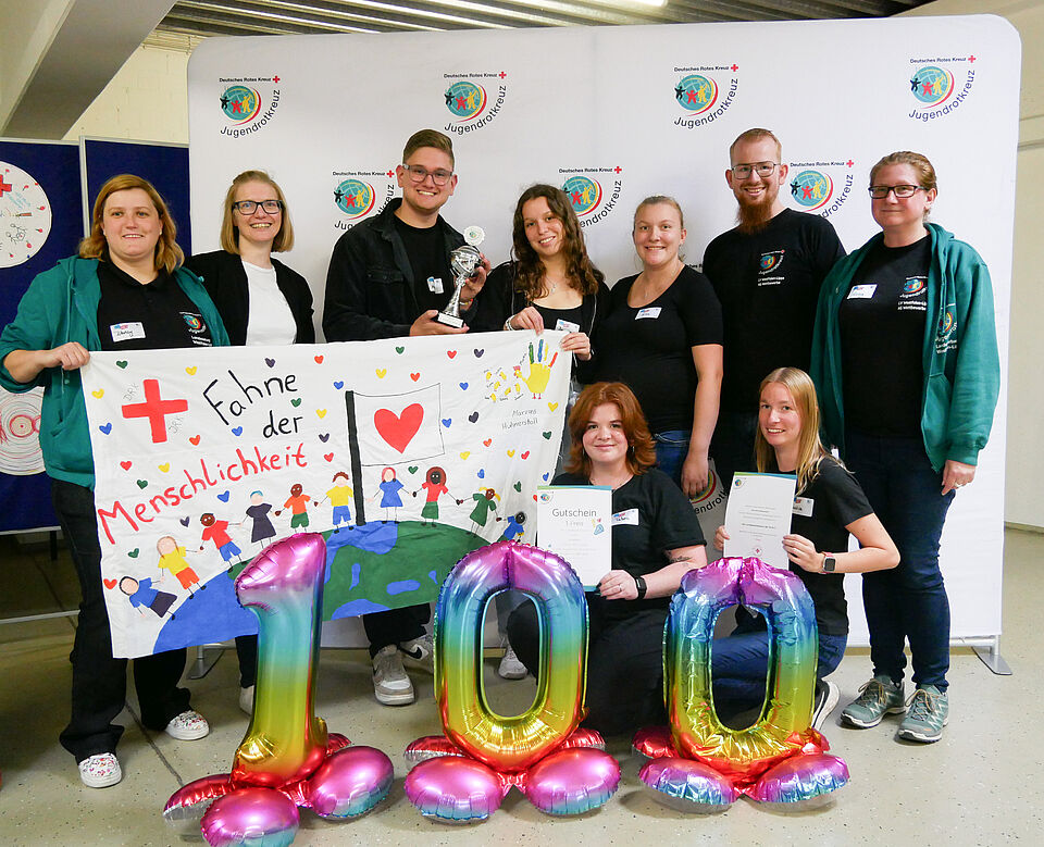 Neun Personen stehen in zwei Reihen vor einer weißen Wand mit dem Logo des Jugendrotkreuzes. Die Gruppe hält ein bemaltes Plakat mit dem Titel „Fahne der Menschlichkeit“, auf dem Figuren und bunte Herzen zu sehen sind. Zwei Personen zeigen Urkunden, eine Person hält einen Pokal. Vor ihnen stehen bunte Luftballons in Form der Zahl 100.