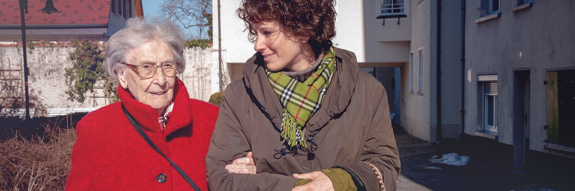 Eine ältere Frau mit Brille und rotem Mantel geht Arm in Arm mit einer jüngeren Frau mit lockigen Haaren und grün-kariertem Schal.