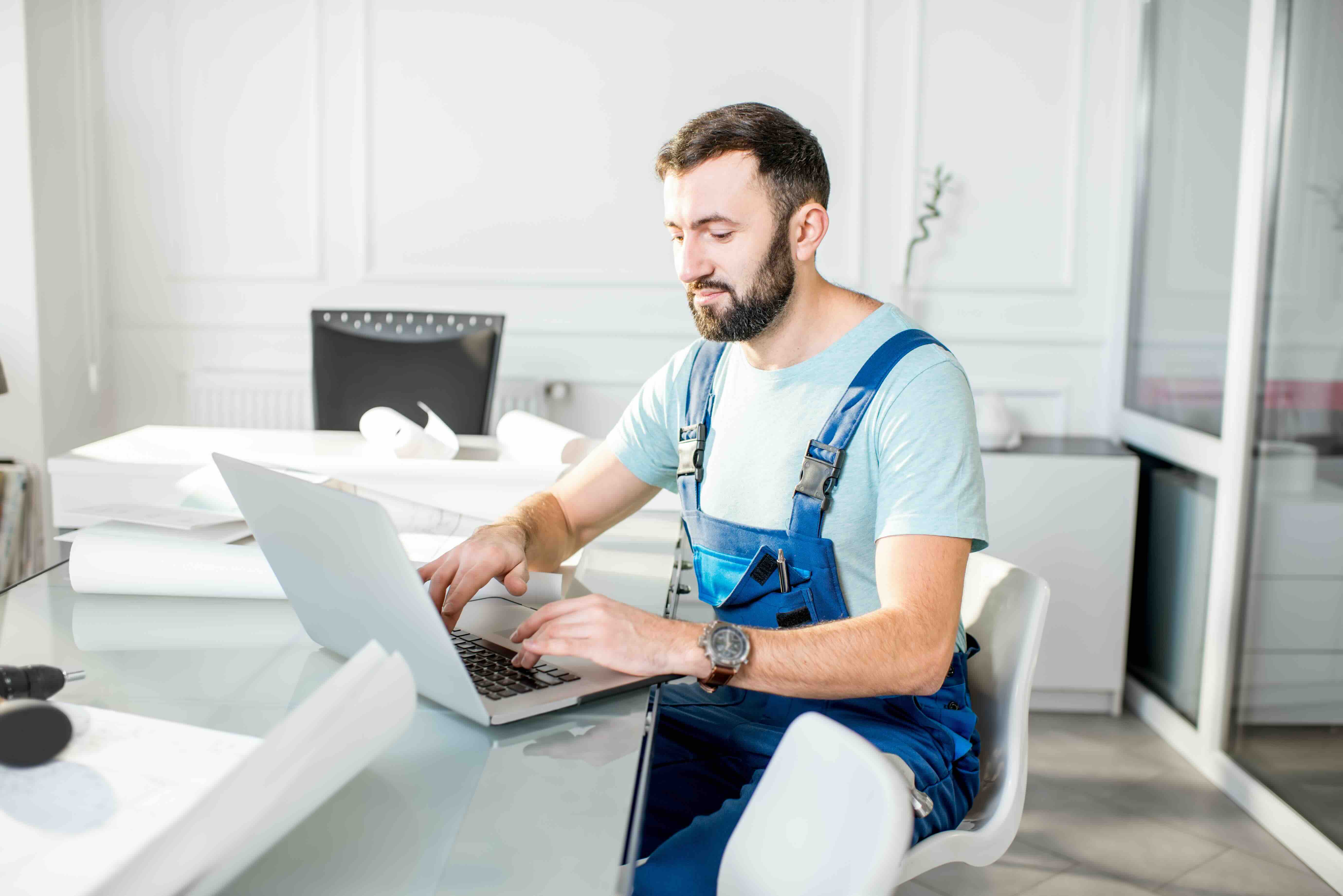 Bartträger in blauer Arbeitskleidung mit Trägern arbeitet an einem Laptop an einem weißen Schreibtisch im hellen Büro.