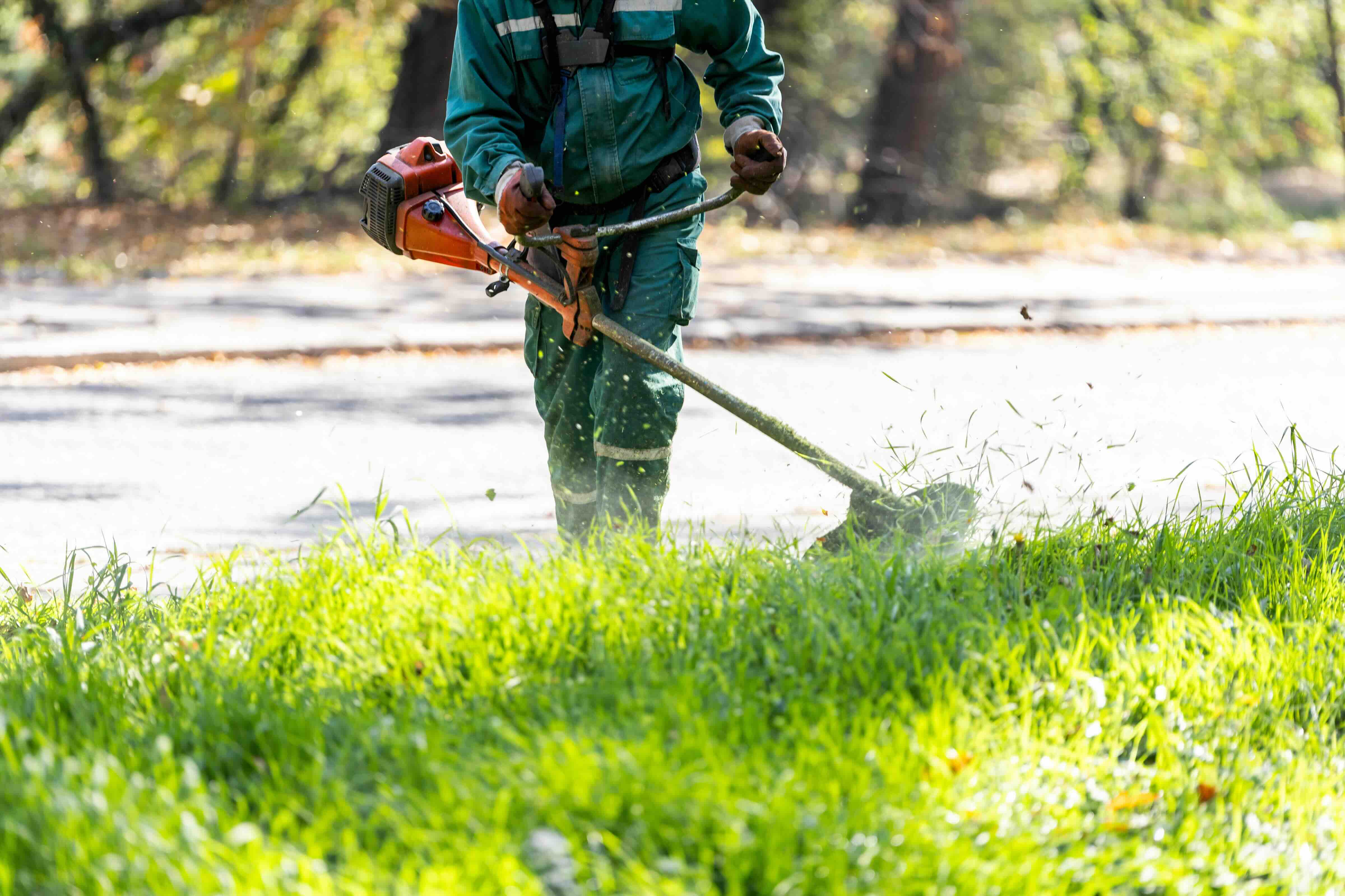 Person in grüner Schutzkleidung mäht mit einer orangefarbenen Motorsense Gras auf einem gepflasterten Weg.