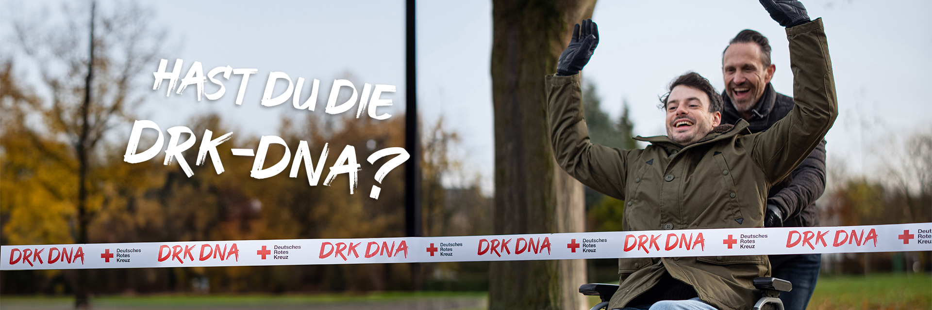 Ein fröhlicher Mann im Rollstuhl mit erhobenen Armen wird von einem anderen lachenden Mann durch ein Zielband geschoben, auf dem "DRK DNA" steht.