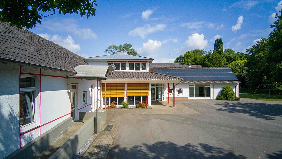 Außenansicht eines weißen Gebäudes mit rotem Rahmen an den Fenstern und gelben Jalousien. Auf dem Dach sind Solarpaneele angebracht, davor liegt eine gepflasterte Fläche.