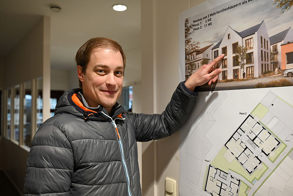 Ein Mann in grauer Steppjacke steht in einem Innenraum und lächelt in die Kamera. Mit einer Hand zeigt er auf ein an der Wand hängendes Poster, das die Visualisierung und den Grundriss zweier geplanter Mehrfamilienhäuser zeigt. Das Poster enthält eine architektonische Außenansicht sowie die Grundrisszeichnung der Gebäude.