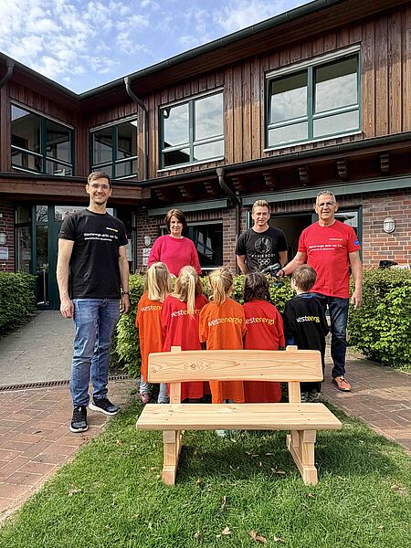 Ein Gruppe von Erwachsenen und Kindern steht vor einem Gebäude mit Holzfassade und großen Fenstern. Die Erwachsenen tragen T-Shirts mit dem Aufdruck „Westenergie aktiv vor Ort“ und halten gemeinsam mit den Kindern, die orange und rote T-Shirts mit dem Aufdruck „Westenergie“ tragen, eine kleine Feier ab. Im Vordergrund steht eine selbstgezimmerte Holzbank, die vermutlich im Rahmen einer Gemeinschaftsaktion entstanden ist. Das Bild vermittelt den Eindruck einer gelungenen Kooperation und eines Gemeinschaftsprojekts.