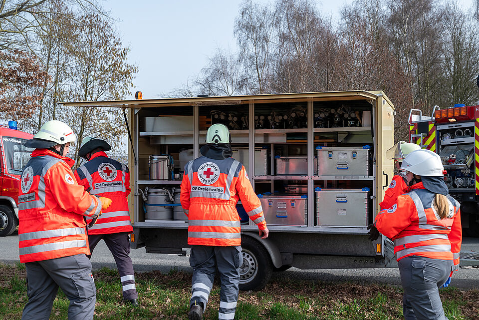 Das Bild zeigt einen Transportwagen des DRK umgeben von Natur. Die Einsatzkräfte tragen Einsatzkleidung und sind mit der Entladung der Materialien beschäftigt. Das heißt, sie laden Verbandsmaterial aus dem Fahrzeug.