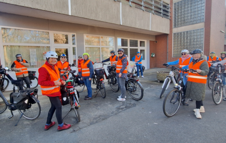 Eine Gruppe von Personen steht im Freien vor einem Gebäude mit großen Fenstern und Backsteinwänden. Alle tragen orange Warnwesten und Fahrradhelme. Sie halten Fahrräder oder stehen direkt neben ihnen. Die Gruppe wirkt vorbereitet für eine gemeinsame Fahrradtour oder eine Veranstaltung.
