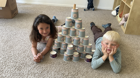  Zwei Kinder, ein Mädchen und ein Junge, liegen auf dem Bauch auf einem beigen Teppichboden. Zwischen ihnen befindet sich ein Turm, der aus vielen grauen Pappbechern gebaut wurde. Das Mädchen auf der linken Seite schaut in die Kamera und lächelt. Der Junge auf der rechten Seite stützt seinen Kopf in seine Hände und schaut ebenfalls in die Kamera. Im Hintergrund sind Regale mit verschiedenen Gegenständen und eine Pappschachtel zu sehen.