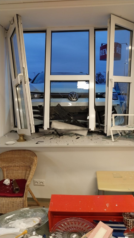 Ein weißes VW-Auto ist durch ein Fenster in ein Gebäude gefahren. Die Scheiben sind zersplittert, Fahrzeugteile und Glasscherben liegen auf der Fensterbank und im Raum. Möbel wie ein Korbstuhl und ein kleiner Tisch stehen zwischen den Trümmern.