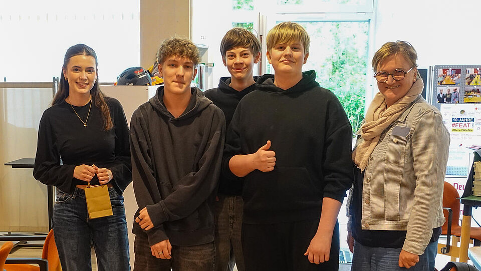 Eine Gruppe von fünf Personen – vier Jugendliche und eine erwachsene Begleitperson – posiert lächelnd für ein Foto in einem hellen Innenraum. Die Jugendlichen tragen lässige Kleidung wie Hoodies und Jeans, eine Person hält eine kleine braune Papiertüte in der Hand. Im Hintergrund sind ein Regal mit Helmen, ein Tisch mit Materialien und ein Plakat .