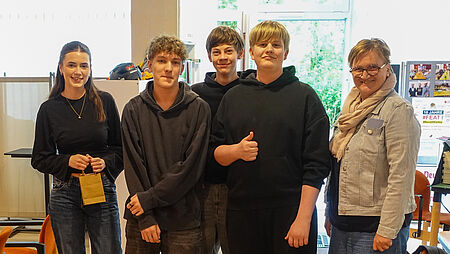 Treffen JRK Wersen Eine Gruppe von fünf Personen – vier Jugendliche und eine erwachsene Begleitperson – posiert lächelnd für ein Foto in einem hellen Innenraum. Die Jugendlichen tragen lässige Kleidung wie Hoodies und Jeans, eine Person hält eine kleine braune Papiertüte in der Hand. Im Hintergrund sind ein Regal mit Helmen, ein Tisch mit Materialien und ein Plakat .