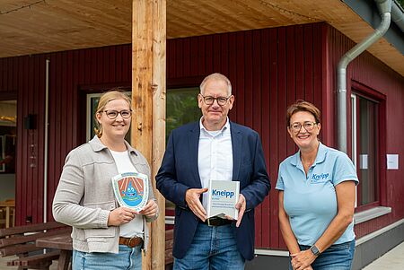 Kneipp-Zertifizierung Drei Personen stehen vor einem roten Holzgebäude mit überdachtem Eingangsbereich. Die Person in der Mitte hält ein Buch mit der Aufschrift „Kneipp Bund“ und trägt ein blaues Hemd mit dem Logo des Kneipp Bundes. Links steht eine Person mit einem Schild, auf dem ein Wassertropfen und ein Regenbogen zu sehen sind. Rechts steht eine Person in einem hellblauen Polohemd mit demselben Logo. Im Hintergrund sind Fenster und eine Holzverkleidung sichtbar.