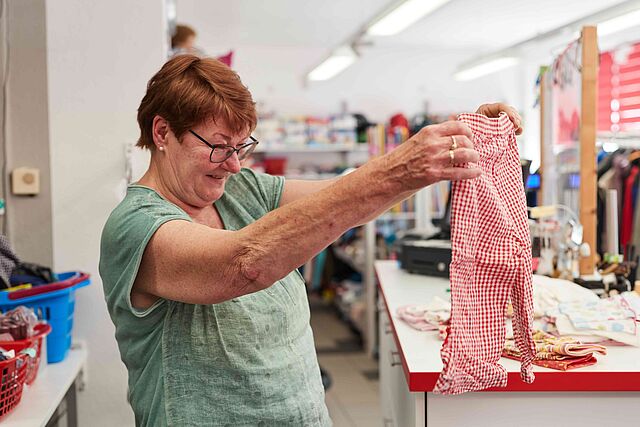 Lachende Frau hält rot-weiß kariertes Kleidungsstück in einem Kleiderladen hoch.