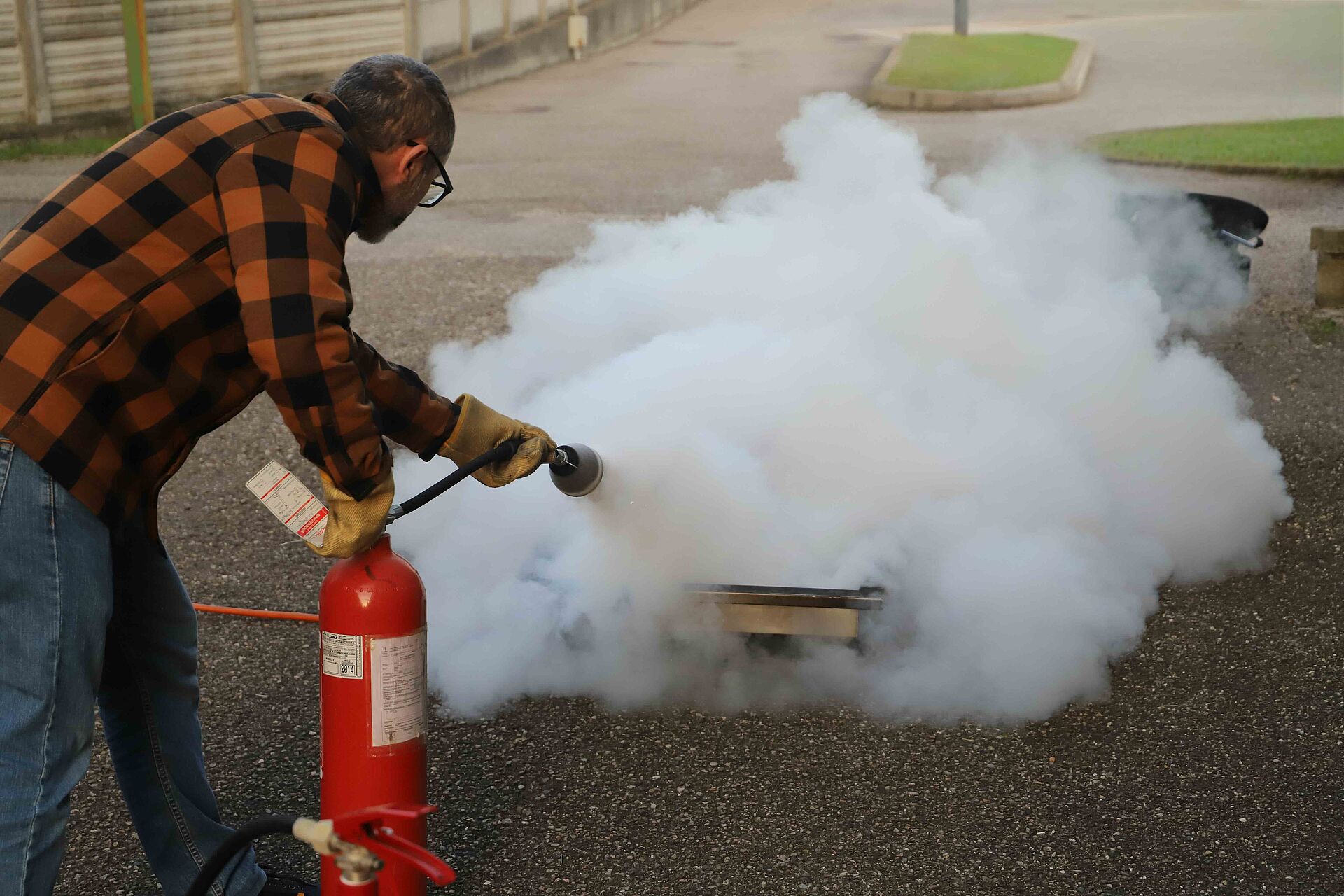 Eine Person löscht bei einer Brandschutzübung im Freien mit einem roten Feuerlöscher einen kleinen Brand am Boden, während dichter weißer Löschnebel aufsteigt.