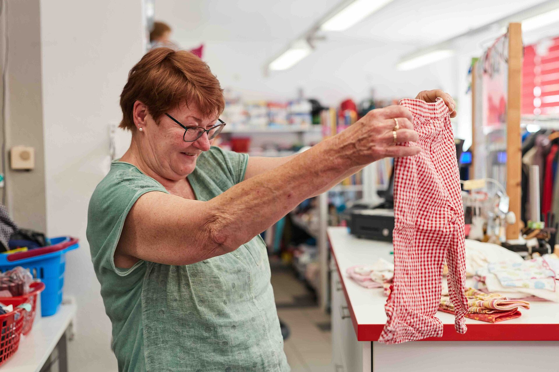 Eine Frau mit Brille und rötlichen Haaren betrachtet lächelnd ein rot-weiß kariertes Kleidungsstück in einem Laden.