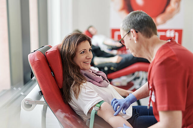 Eine lächelnde Person sitzt in einem roten Blutspendestuhl, während eine medizinische Fachkraft in rotem Shirt mit Handschuhen den Arm der spendenden Person betreut. Im Hintergrund sind weitere Spender zu sehen.