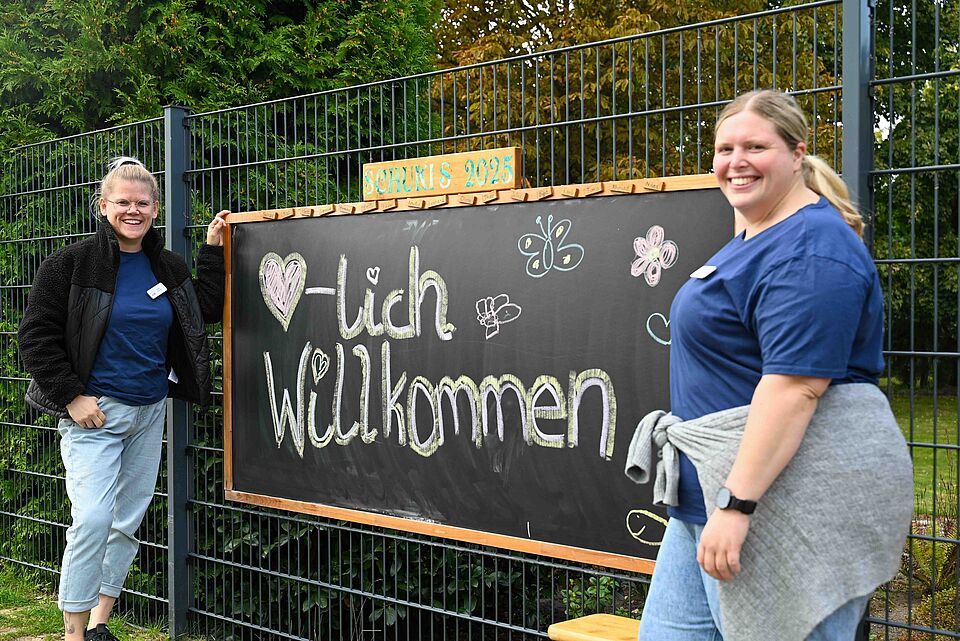 Zwei Mitarbeiterinnen begrüßen Kinder und Eltern neben einer Tafel mit der Aufschrift „Herzlich Willkommen – Schukis 2025“.