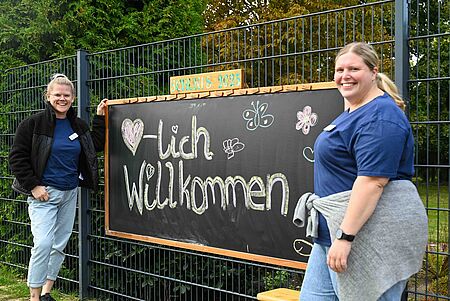 Zwei Mitarbeiterinnen begrüßen Kinder und Eltern neben einer Tafel mit der Aufschrift „Herzlich Willkommen – Schukis 2025“.