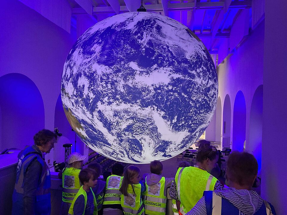 Eine Gruppe von Kindern der Kita 'Schatzkiste' liegt auf dem Boden der Ludwigkirche in Ibbenbüren und bildet einen Kreis. Im Hintergrund schwebt ein großer, beleuchteter Globus der GAIA-Ausstellung, der die Erde zeigt. Die Kinder tragen neonfarbene Sicherheitswesten.