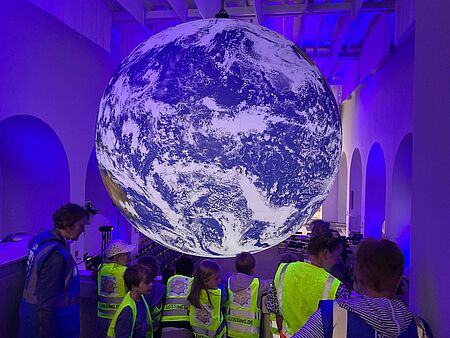 Eine Gruppe von Kindern der Kita 'Schatzkiste' liegt auf dem Boden der Ludwigkirche in Ibbenbüren und bildet einen Kreis. Im Hintergrund schwebt ein großer, beleuchteter Globus der GAIA-Ausstellung, der die Erde zeigt. Die Kinder tragen neonfarbene Sicherheitswesten.
