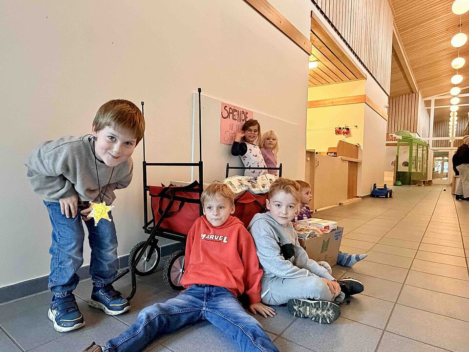 Aktion_Spendensammlung_Schatzkiste Eine Gruppe von Kindern sitzt und steht in einem hellen Flur. Vor ihnen steht ein roter Bollerwagen, daneben ein großer Karton mit gespendeten Lebensmitteln. Hinter dem Wagen hängt ein handgemaltes Schild mit der Aufschrift „Spende für die Tafel“. Die Kinder schauen in Richtung Kamera.