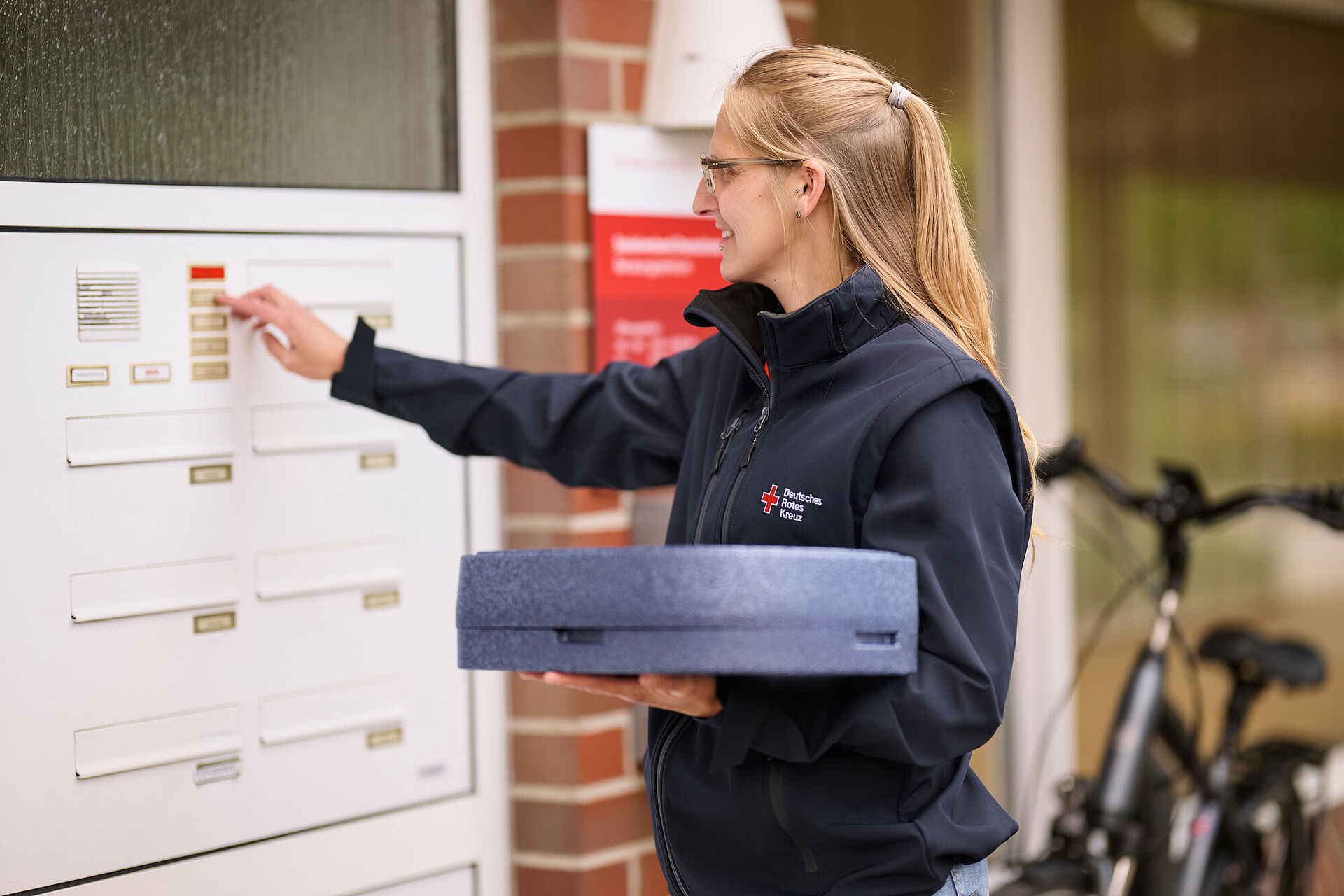 Eine lächelnde Frau in einer DRK-Jacke klingelt mit einer Warmhaltebox in der Hand an einer Haustür.