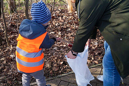 Ein Kind mit Warnweste und Mütze sammelt engagiert Müll in der Natur, begleitet von einer erwachsenen Person, die eine Plastiktüte hält.
