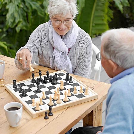 Zwei ältere Personen spielen Schach an einem Holztisch im Freien. Eine Frau mit grauen Haaren und einer hellen Strickjacke zieht eine Schachfigur, während ein Mann mit blauem Hemd zuschaut. Auf dem Tisch stehen zwei Kaffeetassen.