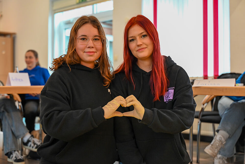 Zwei junge Frauen sitzen nebeneinander, tragen schwarze Hoodies und formen mit behandschuhten Händen gemeinsam ein Herz. Sie blicken in die Kamera, im Hintergrund sind weitere Teilnehmende eines Workshops zu sehen.