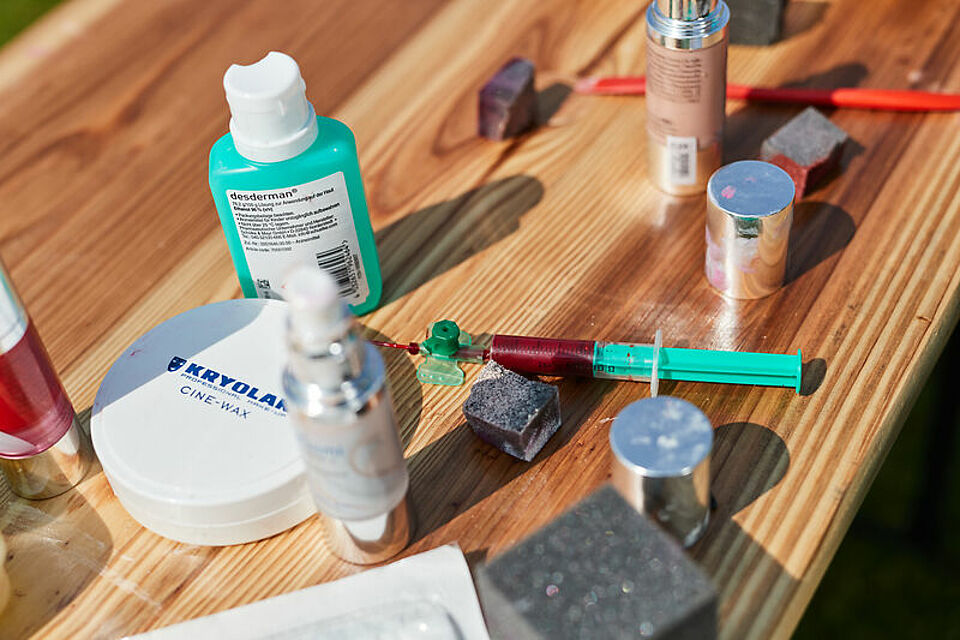 Auf einem Holztisch liegen verschiedene Gegenstände: eine Spritze mit einer grünen Kanüle, eine Tube Desderman-Salbe, ein Behälter mit Kryolan Cine-Wax, ein Make-up-Pinselset, ein rosafarbenes Spray, ein schwarzer Würfel sowie weitere Kosmetik- und Pflegeprodukte.