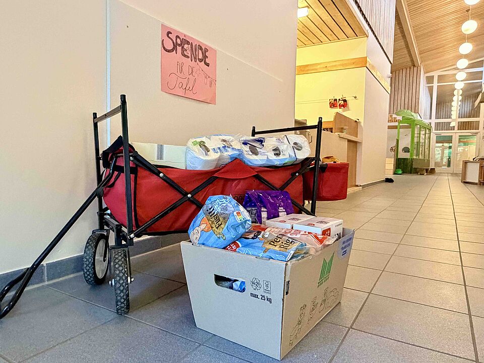 Aktion_Spendensammlung_Schatzkiste Ein roter Bollerwagen und ein großer Karton mit verschiedenen haltbaren Lebensmitteln stehen in einem weitläufigen, hellen Flur. Über dem Wagen hängt ein handgemaltes Schild mit der Aufschrift „Spende für die Tafel“.