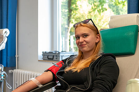 Eine junge Frau sitzt entspannt auf einem Spenderstuhl und lächelt leicht in die Kamera. Sie trägt eine schwarze Jacke und hat eine Blutspende-Nadel im linken Arm, der mit einem roten Halter fixiert ist. Im Hintergrund sind ein Fenster mit grünem Ausblick sowie medizinische Geräte zu sehen.