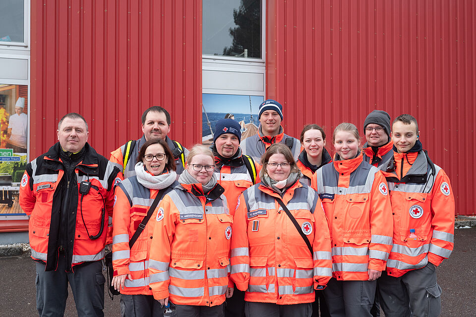 DRK_Karneval Elf Einsatzkräfte des Deutschen Roten Kreuzes stehen in zwei Reihen vor einer roten Gebäudefassade und blicken in die Kamera. Sie tragen orangefarbene Einsatzjacken mit reflektierenden Streifen und DRK-Emblem, teilweise mit Funkgeräten und Mützen. Die Gruppe wirkt geschlossen und einsatzbereit.