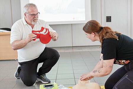 EH-Ausbilder:innen gesucht Ein Mann kniet auf dem Boden und hält eine rote Übungshilfe für die Herz-Lungen-Wiederbelebung (HLW) in den Händen, während eine Frau neben ihm an einer Übungspuppe die HLW durchführt. Beide befinden sich in einem hellen Raum, der für Erste-Hilfe-Schulungen genutzt wird. Im Hintergrund sind ein Tisch und ein AED-Gerät (Automatisierter Externer Defibrillator) zu sehen.