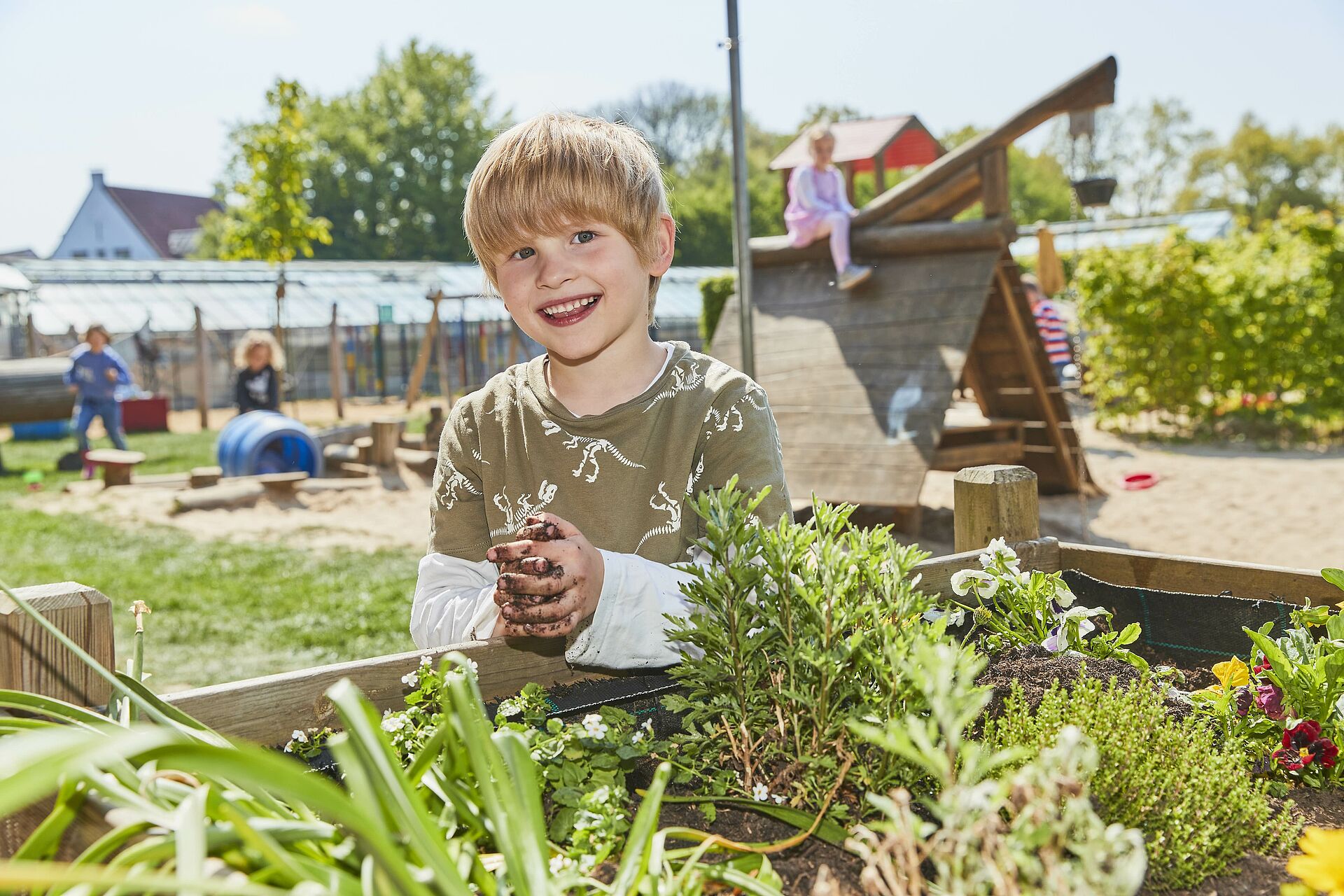 Ein lächelnder Junge mit blonden Haaren und schmutzigen Händen steht vor einem Hochbeet in einem Garten oder auf einem Spielplatz. Im Hintergrund sind weitere Kinder und Spielgeräte zu sehen.
