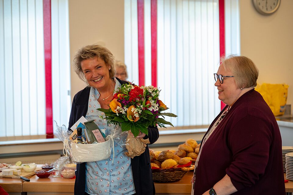 Eine Frau steht lächelnd in einem hellen Raum und hält einen Blumenstrauß sowie ein Geschenkekorb in den Händen. Neben ihr steht eine weitere Frau, die sie freundlich ansieht. Im Hintergrund sind ein Buffet mit Brötchen und Aufschnitt sowie weitere Personen zu sehen. Durch die großen Fenster fällt Tageslicht, rote Vorhänge rahmen den Raum. Die Szene wirkt festlich und herzlich – offenbar ein Anlass zur Gratulation oder Verabschiedung.