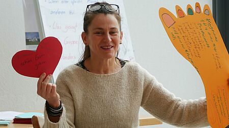 Referentin Eine lächelnde Person in einem hellen Pullover hält in der linken Hand ein rotes Herz aus Pappe mit handschriftlicher Aufschrift und in der rechten Hand eine orangefarbene, großformatige Fußabdruck-Vorlage, die ebenfalls mit handschriftlichen Notizen und Zeichnungen versehen ist. Im Hintergrund sind ein Whiteboard mit Notizen sowie ein Bild an der Wand zu erkennen.