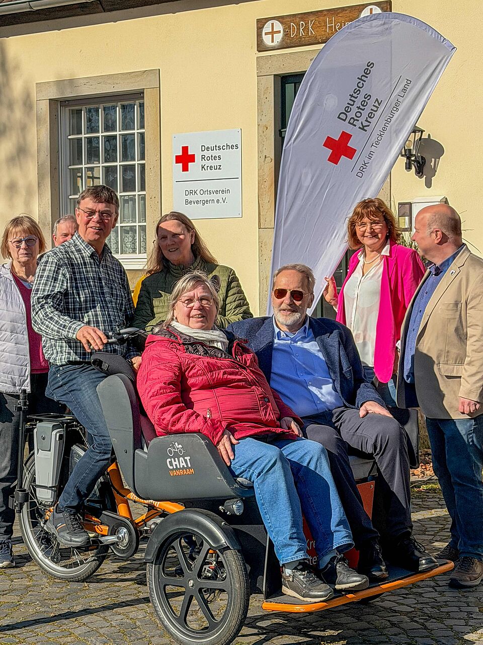 Gruppe von Personen vor dem Vereinsheim des DRK Ortsverein Bevergern e.V., darunter zwei ältere Menschen, die in einer Rikscha des Projekts ‚Radeln ohne Alter‘ sitzen. Im Hintergrund sind das DRK-Schild und eine Flagge des DRK Tecklenburger Land zu sehen.