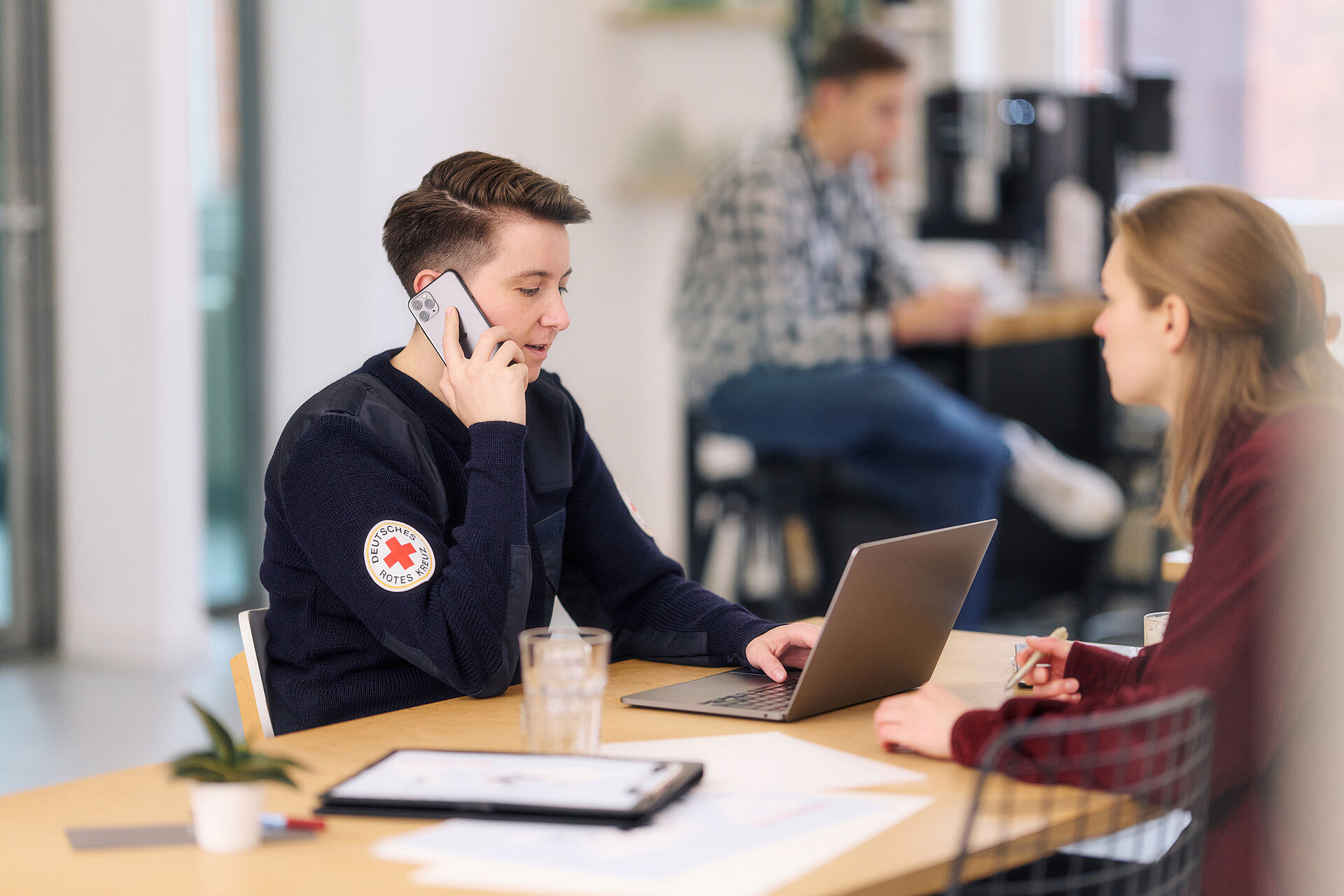 Eine Person in einem dunklen Pullover des Deutschen Roten Kreuzes mit dem Logo am Arm telefoniert und sitzt an einem Tisch mit einem Laptop, während eine weitere Person gegenüber sitzt und Notizen macht. Im Hintergrund ist eine dritte Person unscharf zu erkennen