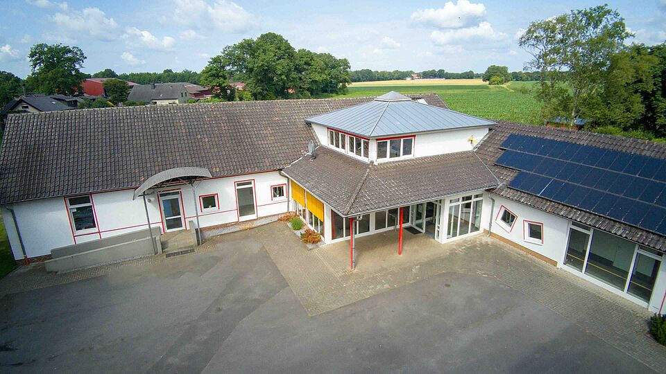 Luftaufnahme eines Gebäudekomplexes mit grauem Dach, weißen Fassaden und Solarpaneelen. Im Hintergrund sind Felder und Bäume zu sehen.