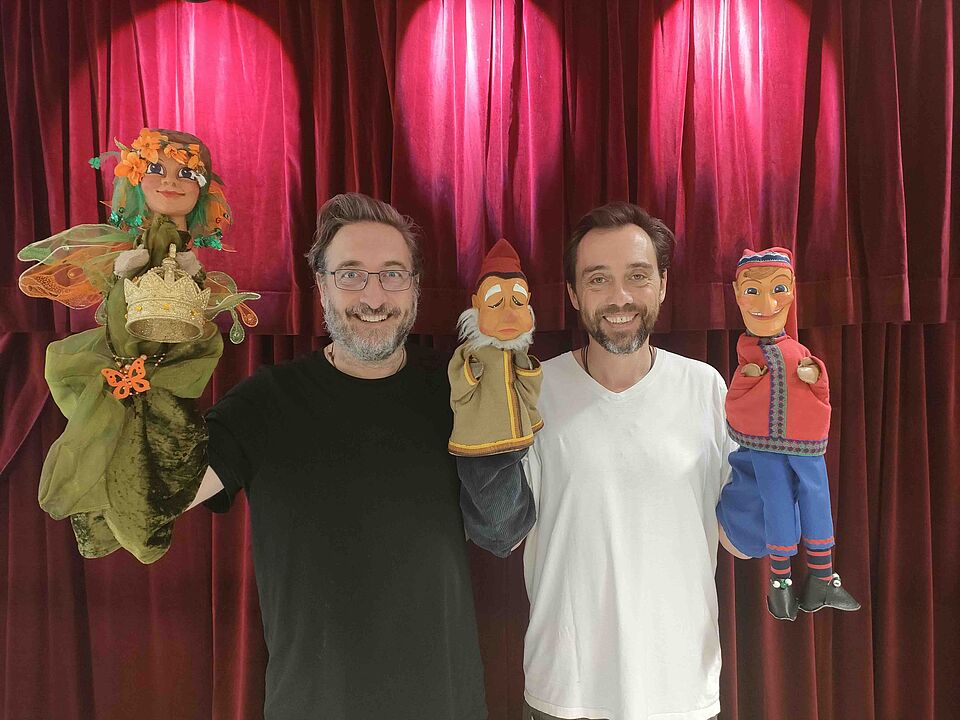Puppenspieler Zwei Männer stehen vor einem roten Samtvorhang und halten jeweils zwei Handpuppen in die Kamera. Der Mann links trägt ein schwarzes T-Shirt und hält eine grüne Fee mit Blumenkranz sowie eine Figur mit roter Mütze. Der Mann rechts trägt ein weißes T-Shirt und hält zwei farbenfrohe Puppen – eine davon in roter Jacke und blauer Hose. Beide lächeln freundlich.