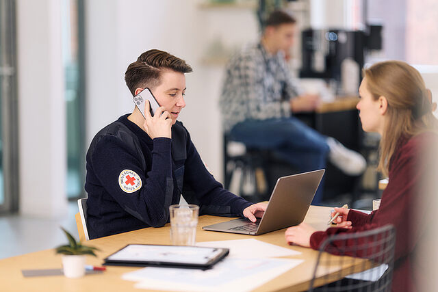 Eine Person in einem dunklen Pullover des Deutschen Roten Kreuzes mit dem Logo am Arm telefoniert und sitzt an einem Tisch mit einem Laptop, während eine weitere Person gegenüber sitzt und Notizen macht. Im Hintergrund ist eine dritte Person unscharf zu erkennen
