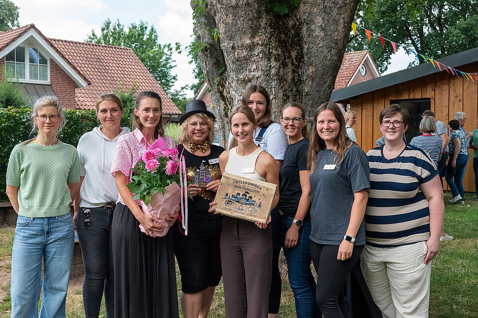 Jubiläum Kita "Burg Funkelstein" Gruppe von neun Personen steht lächelnd im Freien vor einem großen Baum und einem Holzhaus mit rotem Ziegeldach. Eine Person hält einen Blumenstrauß in pinker Verpackung, eine andere ein Holzschild mit der Aufschrift 'Willkommen Burg Funkelstein'. Im Hintergrund sind weitere Menschen, ein Garten und bunte Girlanden zu sehen. Die Stimmung wirkt fröhlich und gesellig