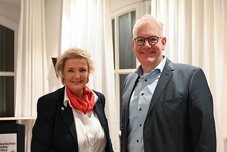 Zwei Personen stehen nebeneinander in einem hellen Innenraum. Links steht eine Frau mit hellem, kurzen Haar, dunkelblazer, weißer Bluse und rotem Halstuch. Rechts steht ein Mann mit kurzem, hellem Haar, Brille und grauem Anzug über einem hellblauen Hemd. Beide lächeln in die Kamera. Im Hintergrund hängen helle Vorhänge und es sind Fenster zu sehen.