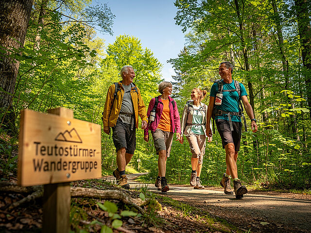 Eine Gruppe von Wandernden genießt einen gemeinsamen Ausflug im Wald. Im Hintergrund und unten rechts ist das Schild der ‚Teutostürmer Wandergruppe‘ des DRK-Ortsvereins Wersen-Büren zu sehen.