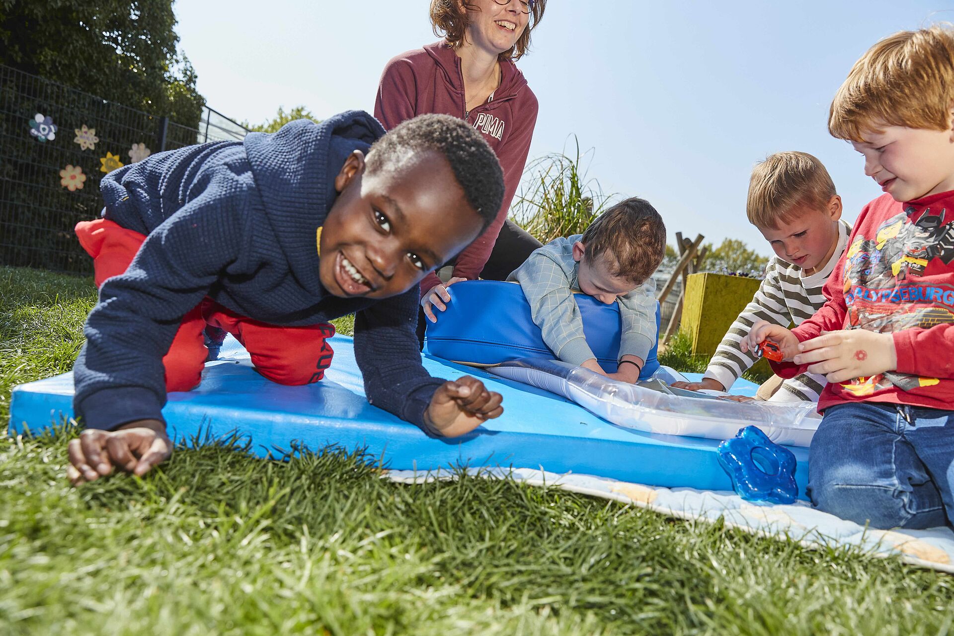 Ein Kind mit dunkler Kleidung lacht in die Kamera, während es auf allen Vieren auf einer blauen Matte im Gras spielt, umgeben von anderen Kindern und einer erwachsenen Person.