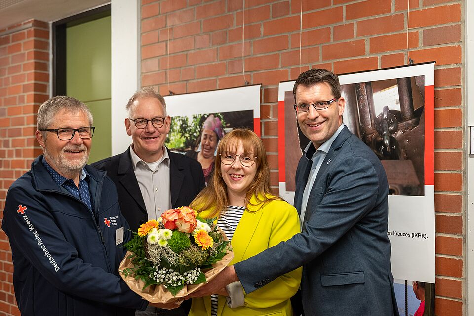 Vier Personen stehen in einem Raum mit Ziegelwänden und Postern im Hintergrund. Eine Frau in gelber Jacke hält einen großen Blumenstrauß in braunem Papier, umgeben von drei Männern in formeller und halbformeller Kleidung. Einer der Männer trägt eine Jacke mit dem Logo des Deutschen Roten Kreuzes.
