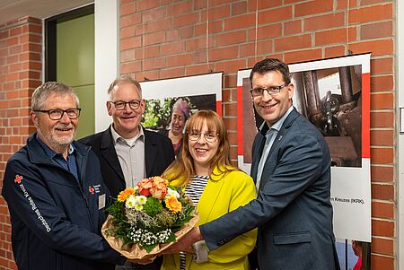 Ausstellung Recke Vier Personen stehen in einem Raum mit Ziegelwänden und Postern im Hintergrund. Eine Frau in gelber Jacke hält einen großen Blumenstrauß in braunem Papier, umgeben von drei Männern in formeller und halbformeller Kleidung. Einer der Männer trägt eine Jacke mit dem Logo des Deutschen Roten Kreuzes.