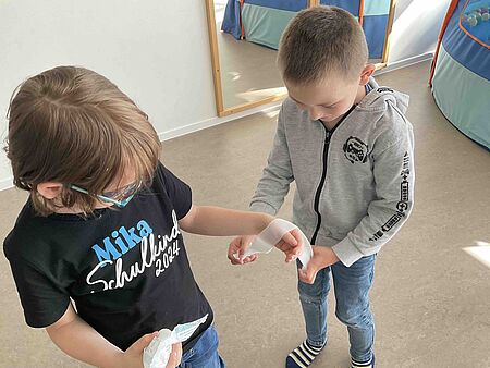 Ein Kind legt dem anderen Kind einen Verband am Arm an. 