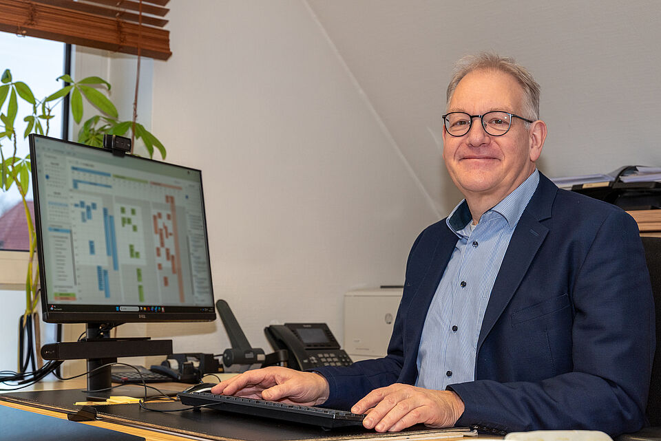 Dr. Friedhelm Höfener sitzt an einem Schreibtisch in einem Büro, blickt lächelnd in die Kamera und arbeitet an einem Computer. Er trägt ein Sakko und Hemd; im Hintergrund sind ein Monitor mit Terminübersicht, Telefon und Büroausstattung zu sehen.